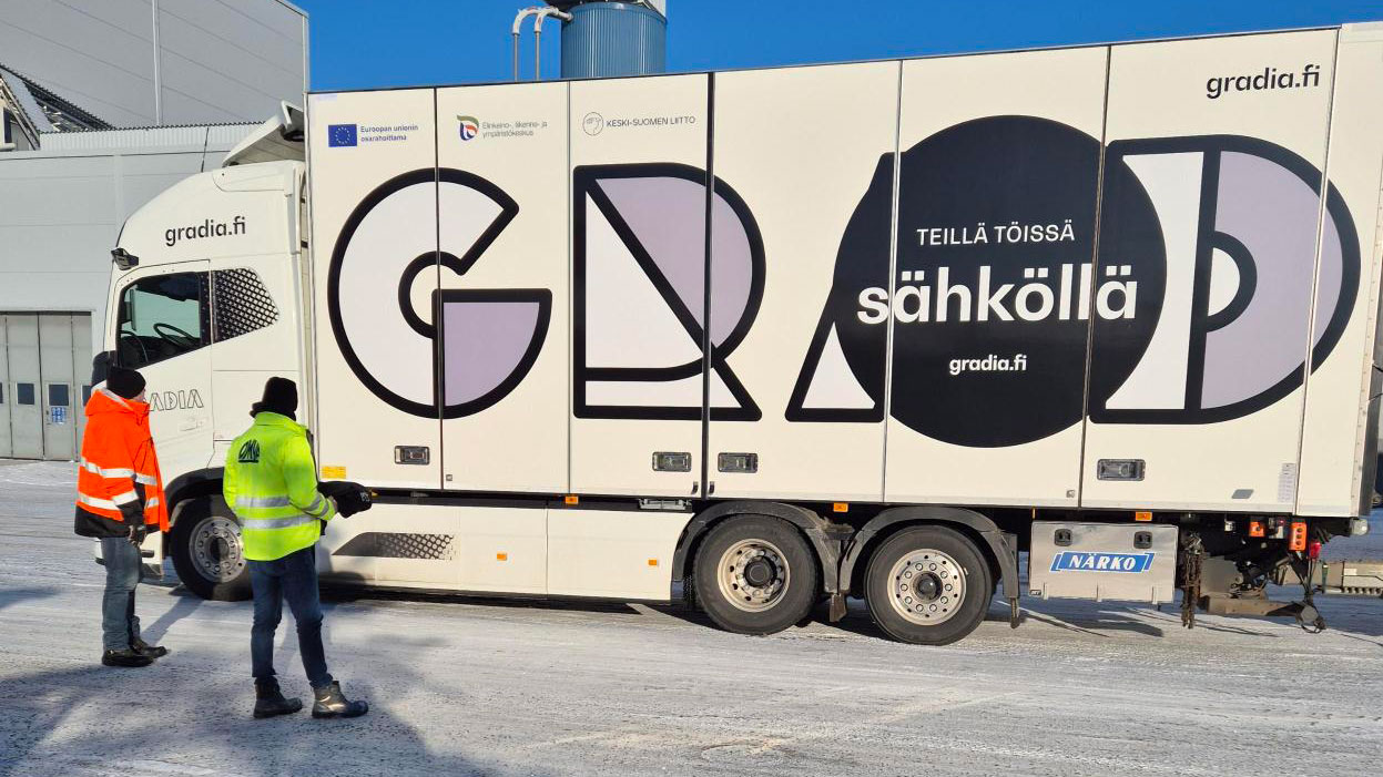 Sähkökuorma-auto talvipäivänä ulkona, edustalla seisoo kaksi henkilöä selin työvaatteissa huomioväreissä.