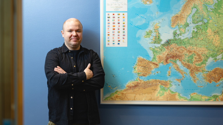 Henkilö seisoo seinällä olevan maailmankartan vieressä ja katsoo kameraan.