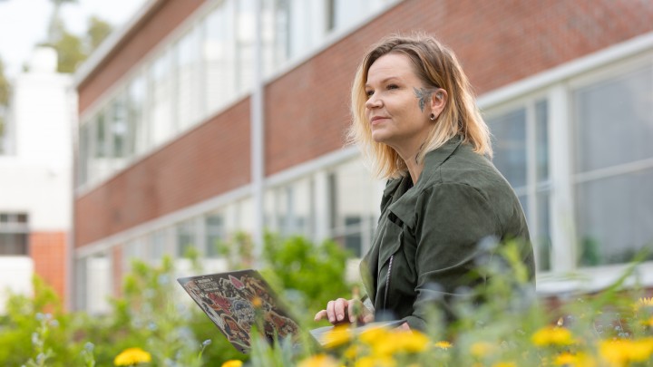 Nainen istuu oppilaitoksen pihamaalla kannettava tietokone kädessään. Oppilaitoksen tiiliseinä taustalla ja etualalla keltaisia voikukkia.