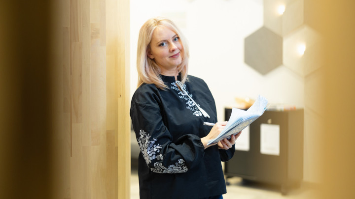 Henkilö pitää kädessään muistivihkoa ja kynää ja katsoo kameraan aulatilassa.