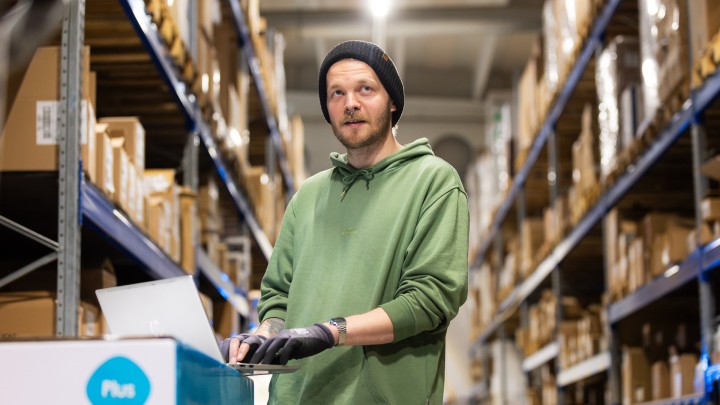 Gradialta valmistunut palvelulogistiikan työtä tekevä Leevi työpaikallaan logistiikkakeskuksessa. Tutkii pakettihyllyjen välissä tietokoneelta toimituksia.