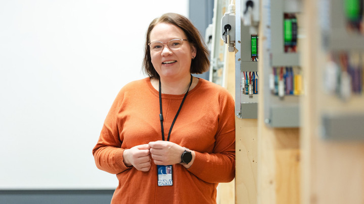 Hymyilevä nainen seisoo sähköalan luokassa sähkökaappien vieressä.